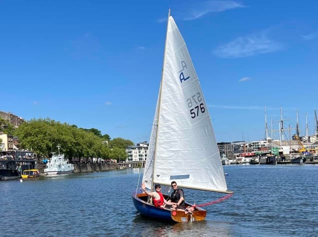 The two SailTies founders sailing a small dinghy across a city harbour on a clear summer day.