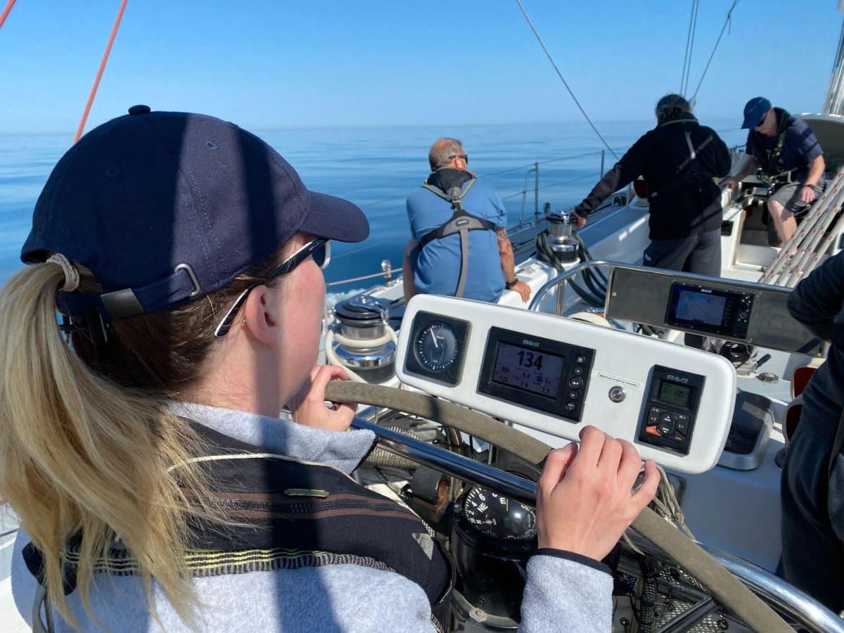 Elouise Pemberton at the wheel offshore in a navy cap and sunglasses, crew working forward on a flat calm sea